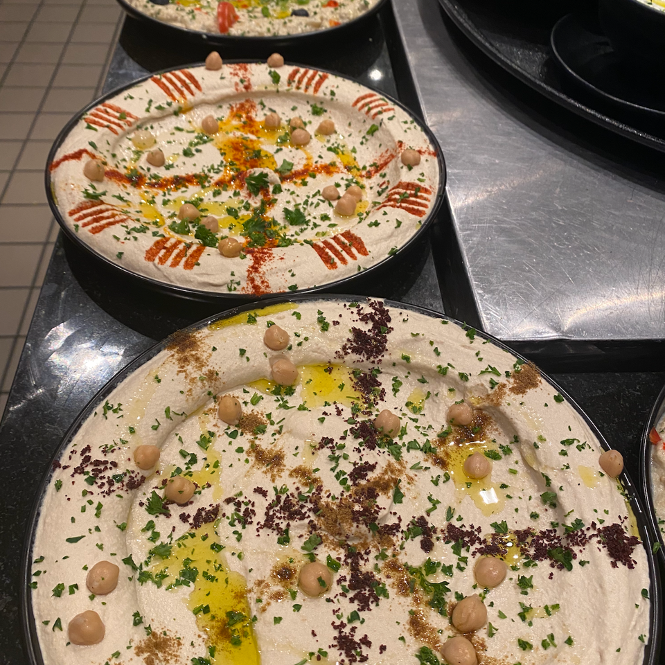 Assorted hummus platters garnished with chickpeas, olive oil, herbs, spices, vegetables, and lemon slices at a buffet.