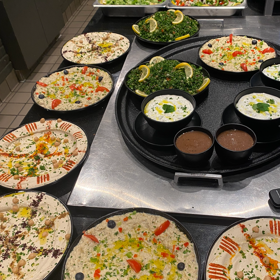 Restaurant buffet displaying hummus, baba ghanoush, tabbouleh, cucumber salad, yogurt dip, and chocolate dessert in black dishes.