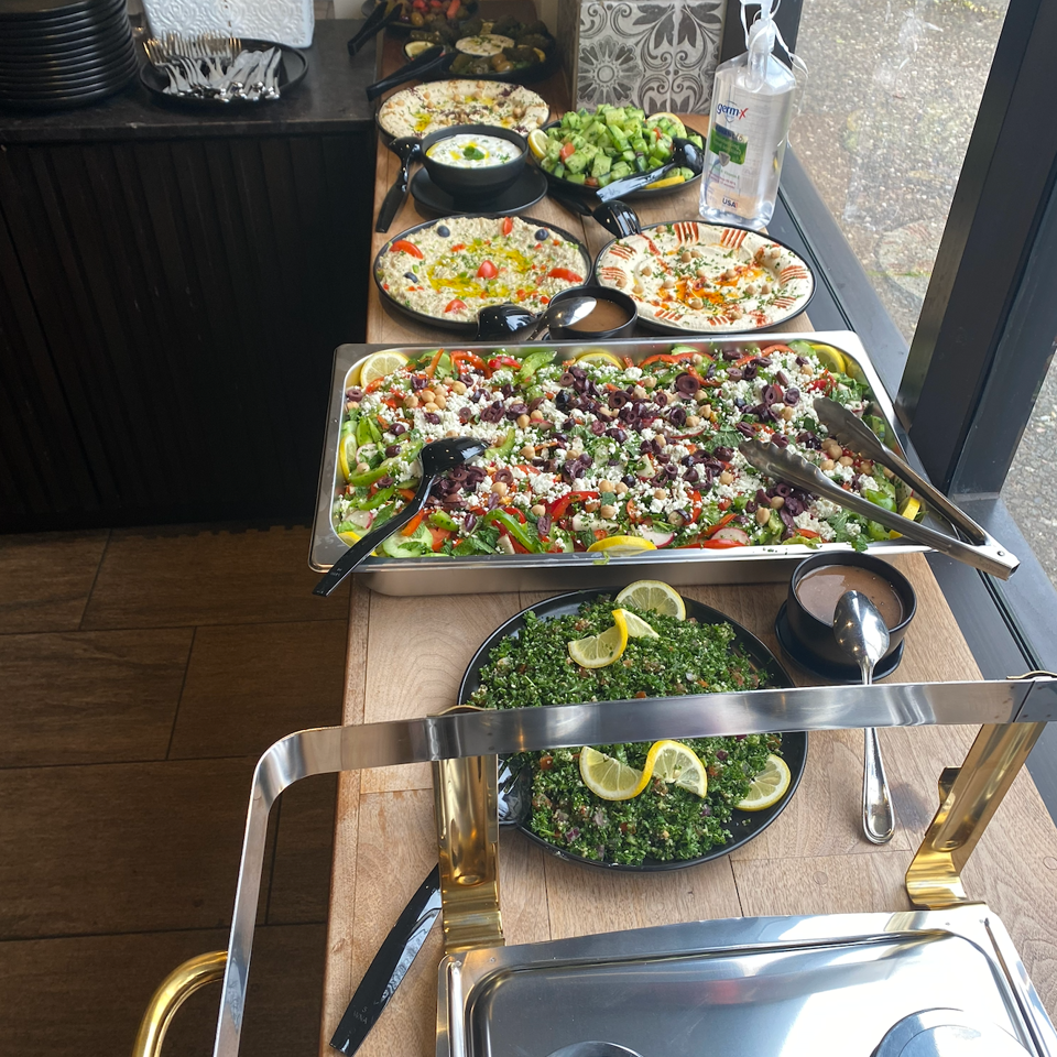 Mediterranean buffet featuring hummus, tabbouleh, Greek salad, vegetables, dips, and serving utensils beside a window.