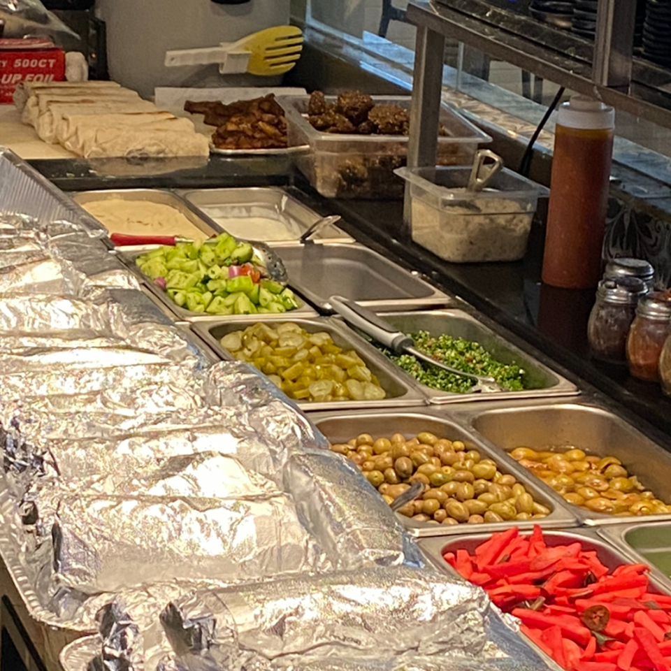 Restaurant counter displaying foil-wrapped food, grilled meats, chopped vegetables, potatoes, pickled peppers, sauces, and serving utensils.