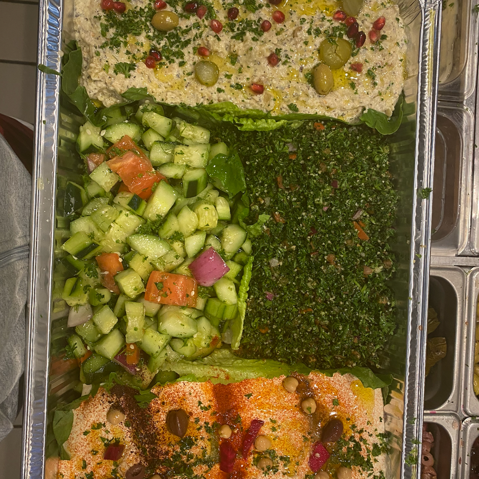 Mediterranean catering platter with hummus, baba ganoush, tabbouleh, cucumber salad, olives, pomegranate, and herbs.